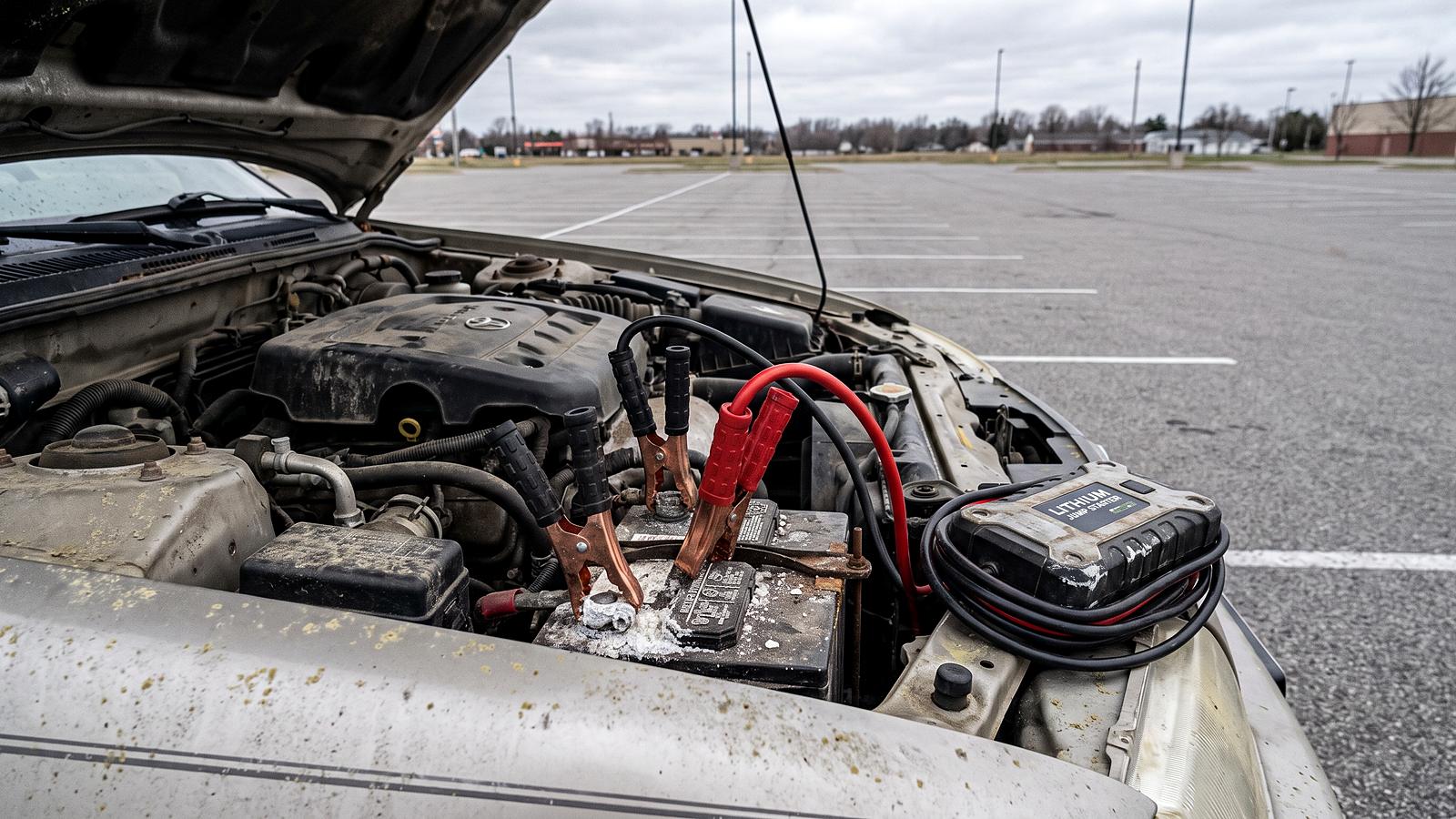 Jumper cable clamps on a car battery terminal under an open hood, a portable jump pack resting alongside